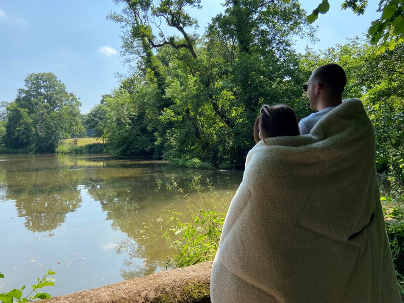 Tonal treetops and vivid spruce Insulated Outdoor Blanko been worn by a young couple enjoying the countryside overlooking a lake with the outdoor blanket, wrapped around them keeping them warm and cosy.