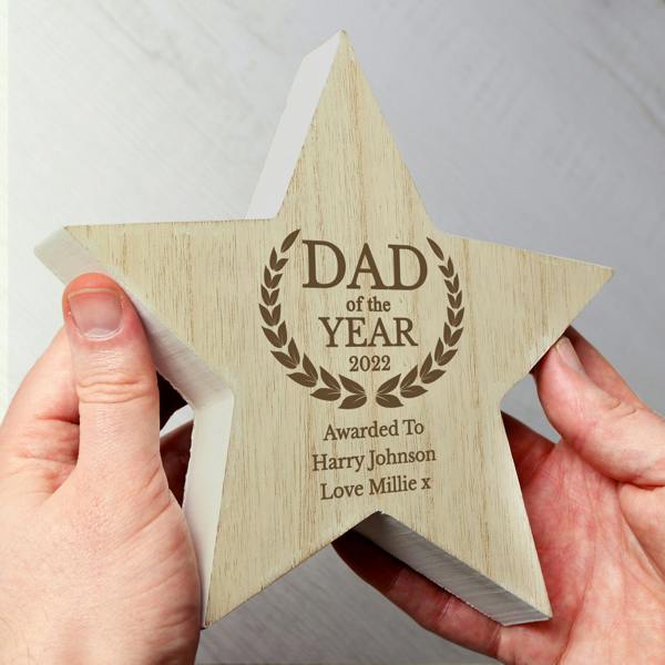 A chunky, free-standing wooden star award being held between two hands. The star features a natural wood grain front with an engraved laurel wreath and the text "DAD of the YEAR 2022," followed by the personalised message "Awarded To Harry Johnson Love Millie x."