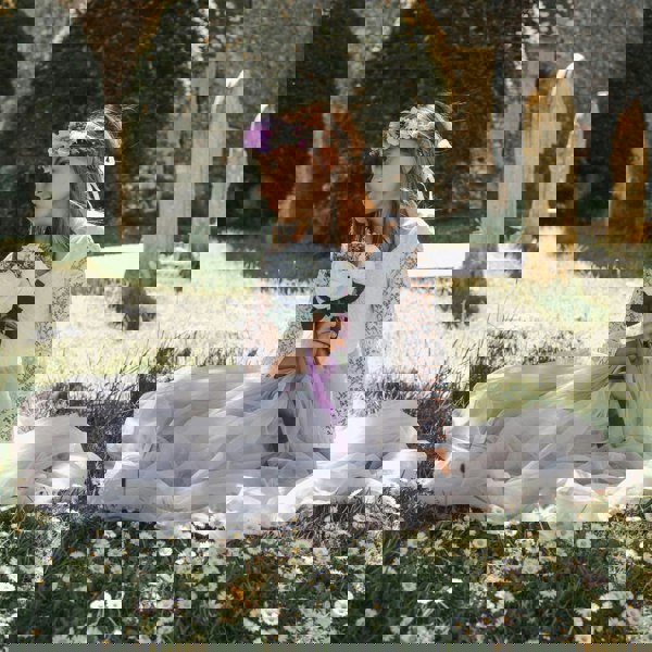 Young girl in a white dress with a floral headband sitting on grass with flowers.