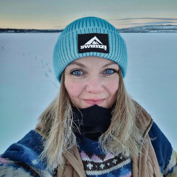 Woman wearing a simple beanie hat knitted in a chunky single rib in a sky blue shade against the snowy backdrop. There is a SWIMZI badge sewn to the front of the deep turn back headband.