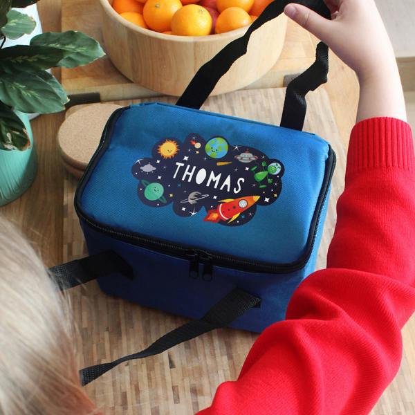 A child wearing a red jumper holds the strap of a personalised blue space lunch bag printed with the name Thomas.