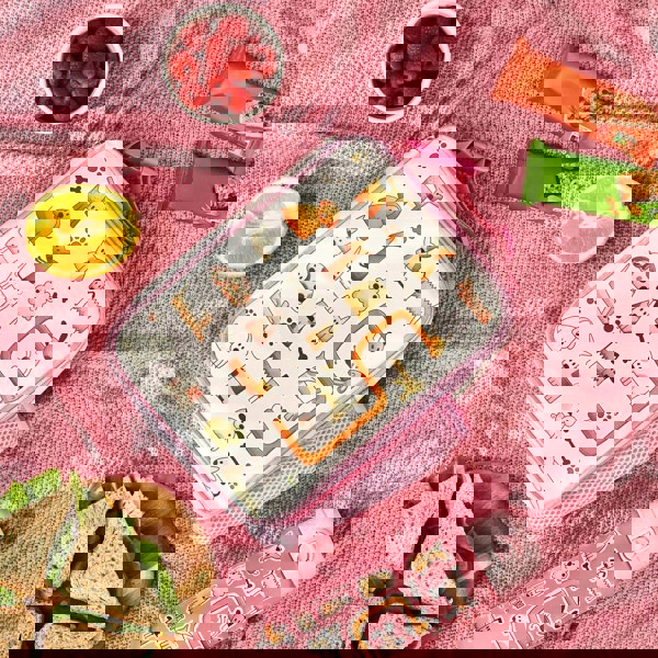 Lunchbox decorated with cartoon dogs is resting on a pink blanket, accompanied by a bowl of raspberries, orange half, sandwich plate, two snack bars, and a dog-themed bottle.