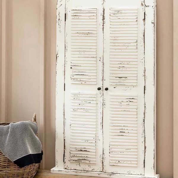 The Château white arched window mirror with fully closed shutter doors leaning against a soft beige wall next to a wicker basket.