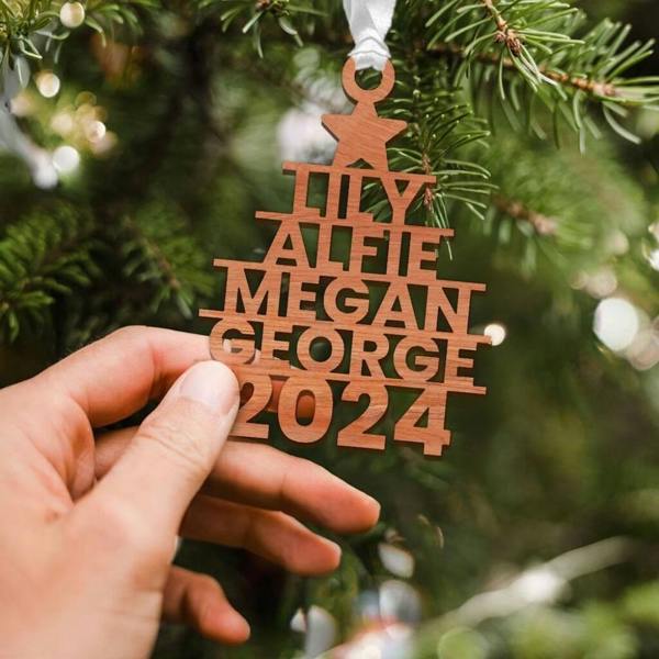 A hand holding a wooden Christmas tree decoration carved with the names 'LILY, ALFIE, MEGAN, GEORGE' and the year '2024' below a star, hanging on a pine tree branch with a white ribbon.