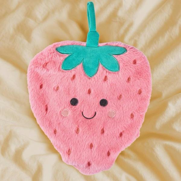 An adorable plush pink Strawberry Hot Water Bottle with a smiling face and green stem resting on a cream bed sheet.