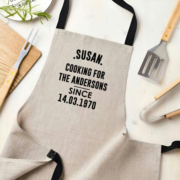 A neutral linen kitchen apron flat lay, custom printed with 'Susan, Cooking for the Andersons Since 14.03.1970' alongside wooden cooking utensils and a carving fork.