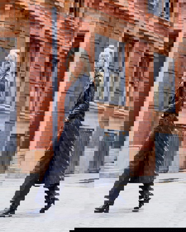 Image features a woman striding across the frame wearing a black leather trench coat.