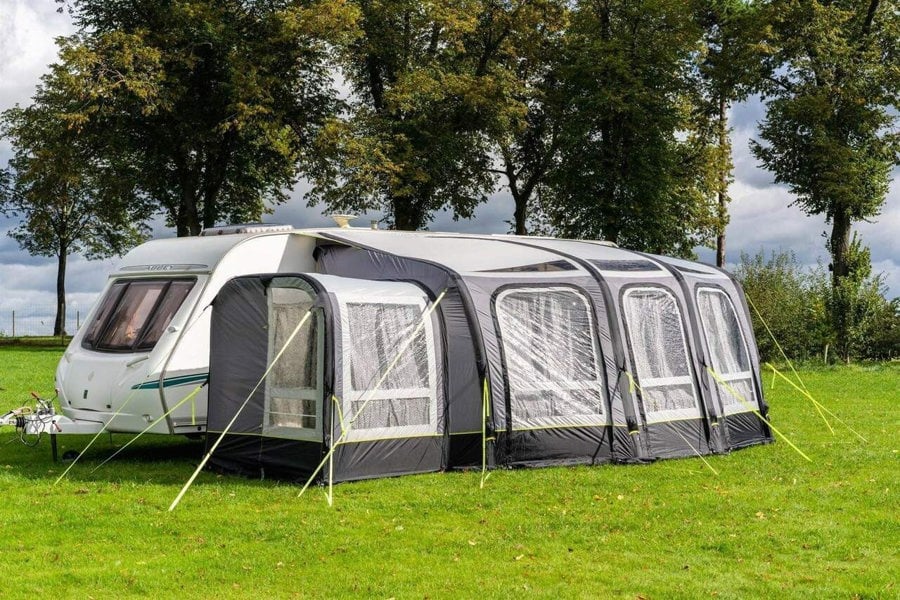 The OLPRO View 420 Caravan Inflatable Porch Awning With Porch Extension attached to a caravan at a campsite with a treeline in the background.