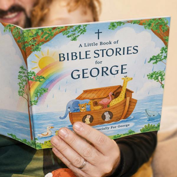 A close-up lifestyle photograph of an adult holding open the front and back covers of the Personalised Children's Bible Stories Book from Abode Home & Gifts custom printed for George, showcasing a watercolour illustration of Noah's Ark under a rainbow.