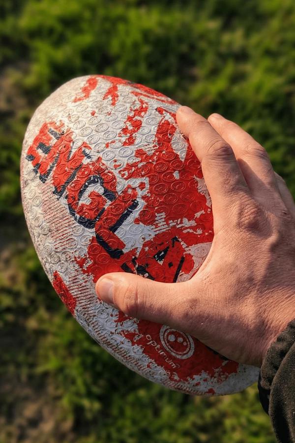 Hand holding a white and red Optimum England rugby ball with ENGLAND print above grass - Optimum