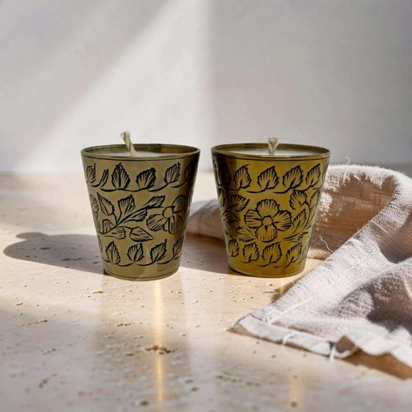 Two small metal candle holders with a gold finish and black floral line patterns, shown on a light stone surface with a folded beige cloth beside them.