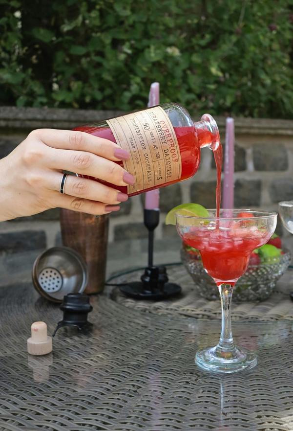 A hand pours an overproof strawberry daiquiri from Prohibition Cocktails from a bottle with a tan paper label into a coupe glass on an outdoor patio table.