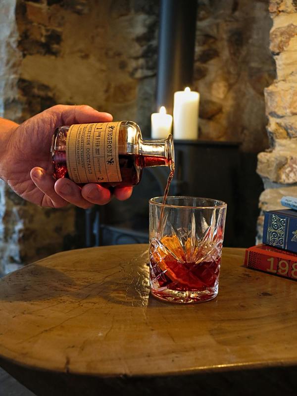 A hand pours a dark amber liquid - Negroni by Prohibition Cocktails - from a small bottle with a cream label into a cut-glass tumbler on a rustic wooden table, with candles and stone walls in the background.