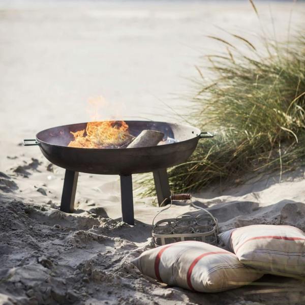 Medium 75cm cast iron garden fire bowl with a lit fire, styled on a sandy beach at sunset with outdoor cushions and a drink carrier nearby.
