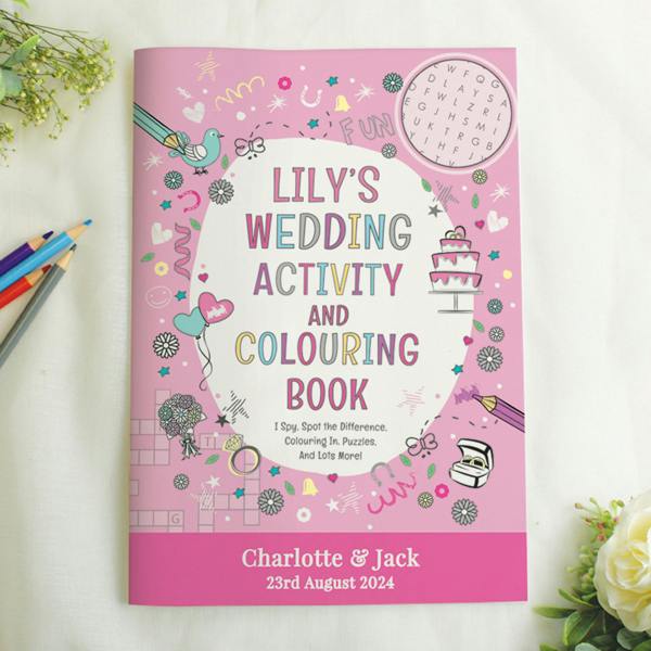 A personalised pink wedding activity and colouring book sits flat on a white fabric background. The cover reads Lily's Wedding Activity and Colouring Book, with a lower pink banner customized with Charlotte and Jack, 23rd August 2024. Colouring pencils and white flowers frame the book.