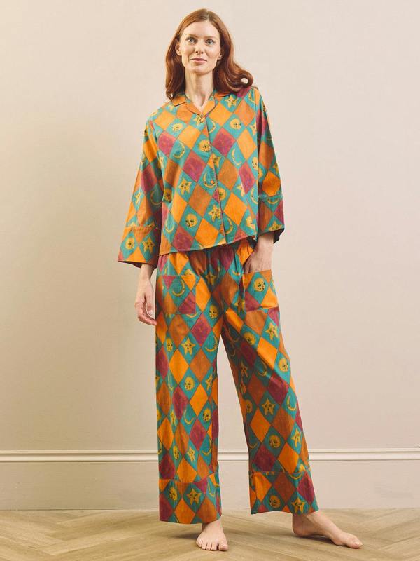A woman stands barefoot indoors wearing a matching two-piece pyjama set with a bold geometric harlequin pattern in orange, teal, burgundy, and yellow. The set includes a long-sleeve button-front top and wide-leg pants.