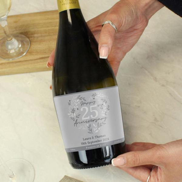 A close-up of a person holding a bottle of personalised 25th Silver Wedding Anniversary Prosecco with a custom grey label featuring a botanical heart motif and custom text reading Laura and Thomas 19th September 2024.