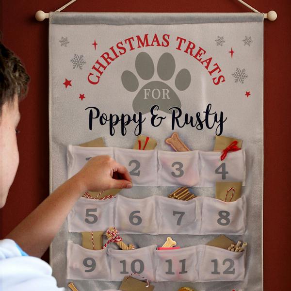 A hand reaching into the pockets of a personalised grey fabric pet advent calendar reading "Poppy & Rusty" hanging on a dark red wall.