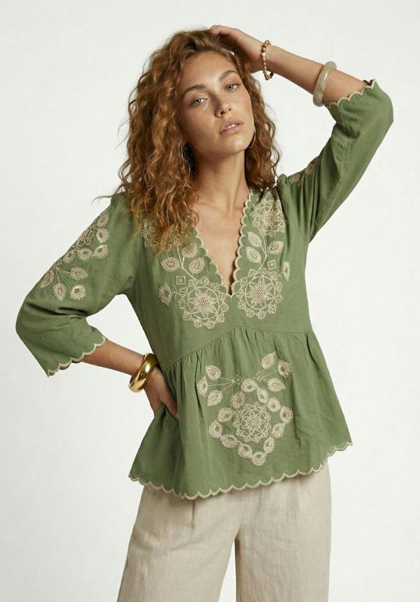 Woman wearing a green embroidered blouse with beige pants on a white background