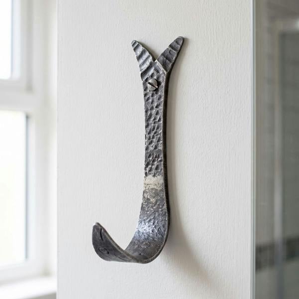 Close-up of hand-forged antique iron fish design coat hook mounted on painted bathroom wall, showcasing craftsmanship and textured surface with natural light.