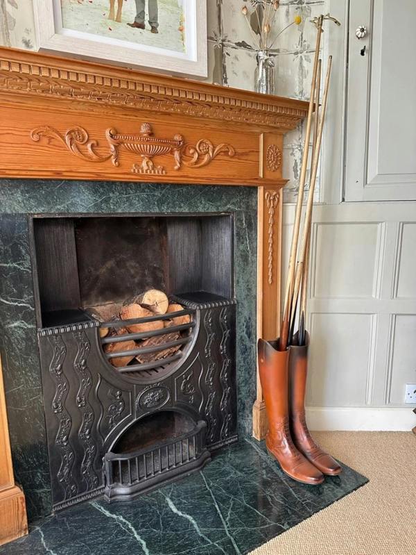 A brown faux-leather riding boot umbrella stand holding vintage walking sticks, styled on a dark green marble hearth next to an antique cast iron fireplace with a wooden mantelpiece.