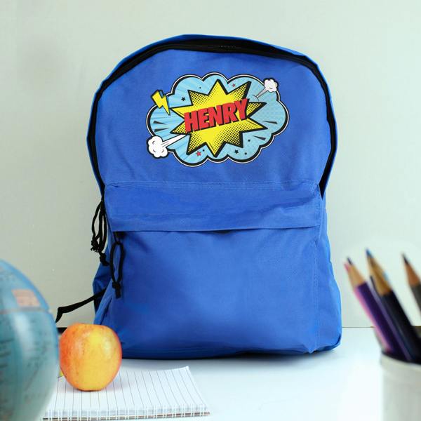 A personalised blue backpack printed with the name HENRY, styled on a desk alongside an apple, a notebook, a globe, and coloured pencils.