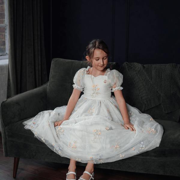 Young girl in a white dress with floral embroidery sitting on a dark green couch.