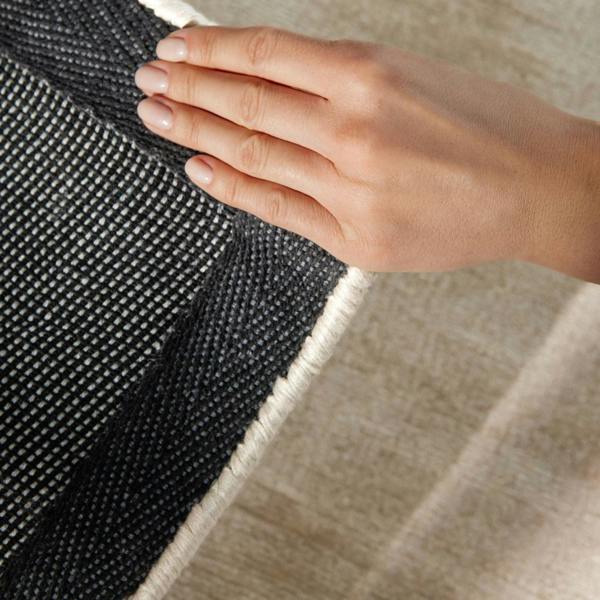 Hand touching the edge of a black and white textured rug on a beige floor.