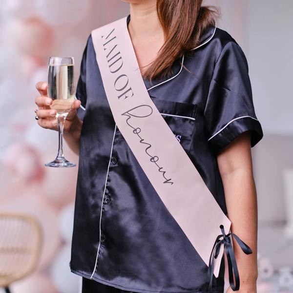 Mid-section view of a woman wearing a pale pink Maid of Honour sash with black text over a black satin pyjama shirt while holding a champagne glass.