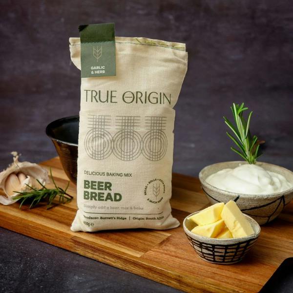 A freshly baked golden beer bread loaf in a lined tin sitting on a wooden board next to a sack of Garlic and Herb Beer Bread mix, fresh rosemary, and a garlic bulb.
