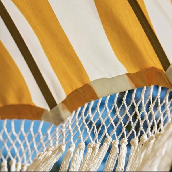 An angled close-up view from beneath the Sunningdale garden parasol looking out towards a soft blue sky. The shot details the yellow and cream striped canvas canopy edge, showcasing the premium knotted cotton macrame mesh netting and long hanging tassel trim.
