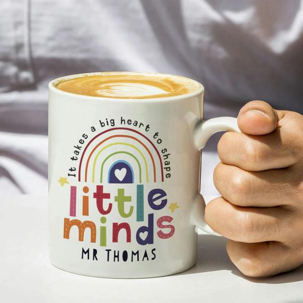 A white ceramic mug featuring a colourful rainbow design and the text 'It takes a big heart to shape little minds MR THOMAS' in uppercase text, held by a person.