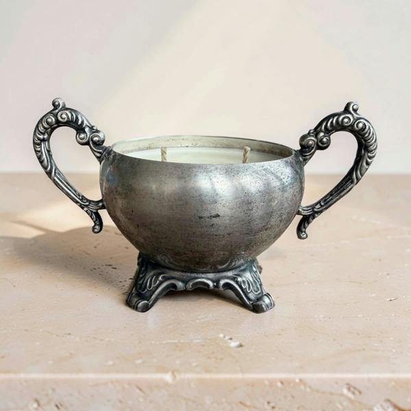 An ornate silver-toned metal candle holder with two decorative handles and a pedestal base, shown filled with white candles on a light beige stone surface against a soft beige background.