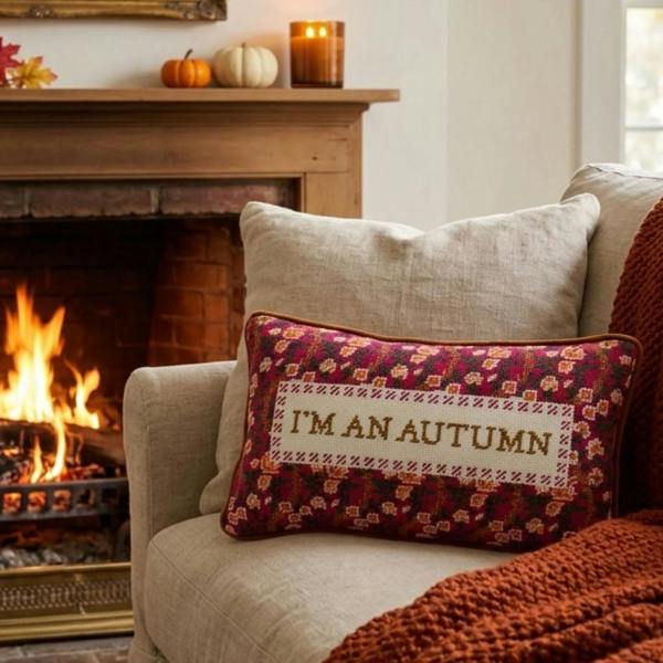 The 'I'm An Autumn' needlepoint statement cushion resting on a neutral linen armchair next to an orange textured knit throw, positioned in front of a rustic brick fireplace with a roaring fire and seasonal mantel decorations.