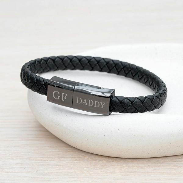 A close-up view of the black woven leather men's tech bracelet with its polished gunmetal clasp engraved with GF and DADDY resting on a white stone dish.
