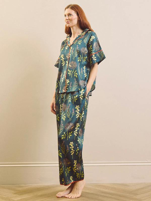 A woman stands barefoot in a studio wearing a matching short-sleeve button-front top and wide-leg pants pyjama set in a dark teal-blue colour with a botanical print in green, yellow, and orange.