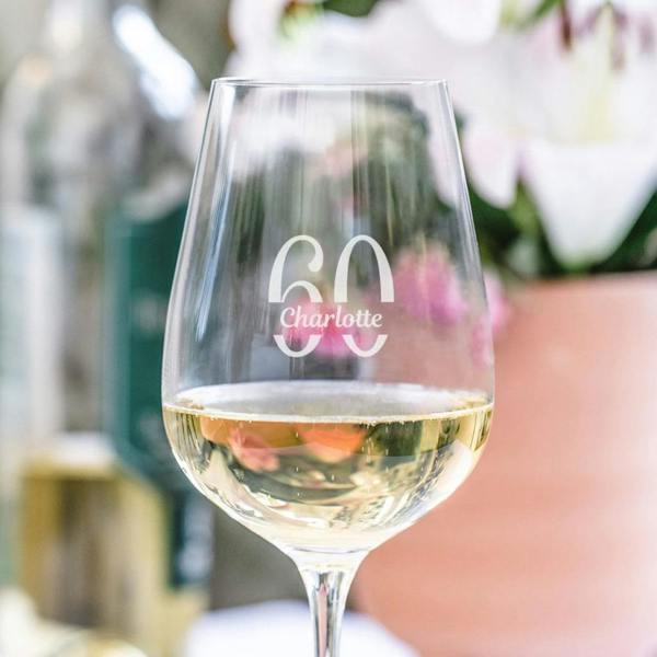A clear wine glass filled with white wine, engraved with the large milestone age '60' above the name 'Charlotte' in clean, white lettering, set against a soft, blurred background.