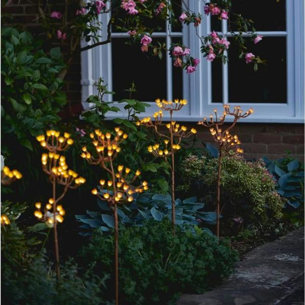 An illuminated solar and battery wild fennel branch stake glowing with 40 warm white LEDs at twilight in a lush cottage garden border.