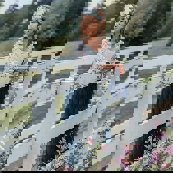 Young girl in a white lace top and blue skirt standing on a white wooden bridge with greenery in the background.