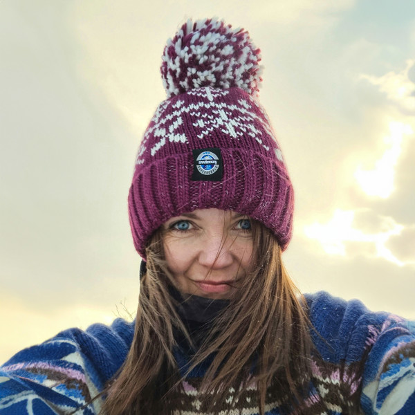  Woman sitting on the snow of Northern Norway is wearing a chunky Nordic knit bobble hat. The burgundy red base and white Nordic design hat also has a deep ribbed headband, contains reflective yarns and has a luxury sherpa fleece lining.