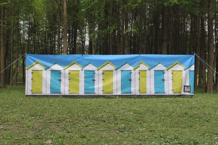 Beach Huts Design 4 Pole Compact Windbreak Steel Poles by OLPRO pictured on a field. It's Blue sky with White Beach huts with alternate blue and yellow doors fully assembled in a straight line.