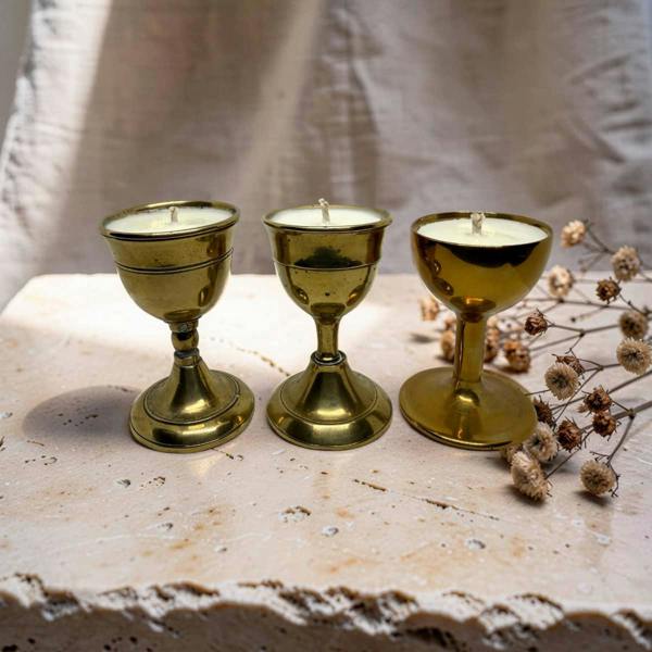Three brass candle holders with white candles, arranged on a rustic stone surface next to dried flowers against a draped fabric backdrop.