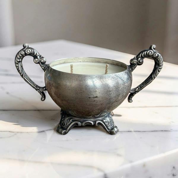 An ornate silver-toned metal candle holder with two decorative handles and a footed base, filled with white wax, set on a light marble surface.
