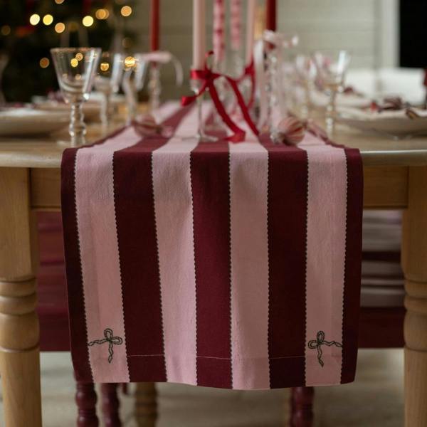 A close-up shot focusing on the end of a burgundy and pink striped organic cotton table runner hanging off a table, highlighting the dark green embroidered bows on the corners.