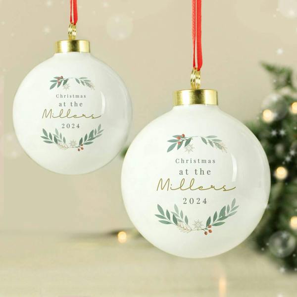 Two glossy white Personalised Christmas Foliage Baubles reading Christmas at the Millers 2024, hanging from red ribbons next to a festive pine tree.