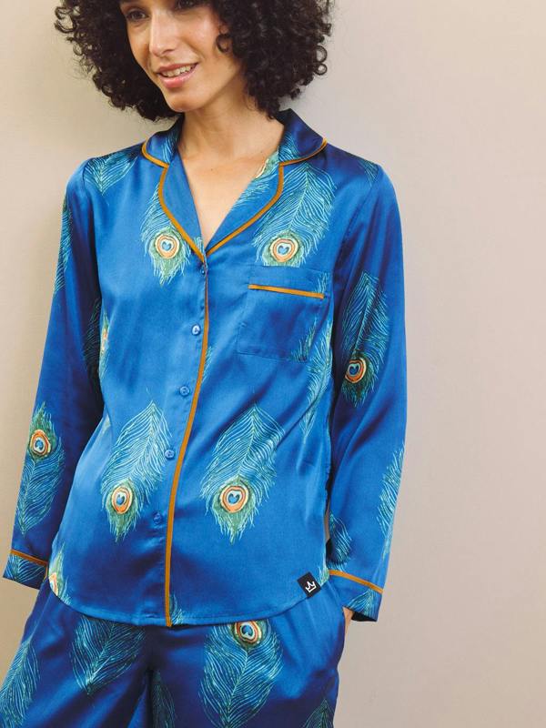 Woman wearing a blue satin pajama set with peacock feather design against a beige background