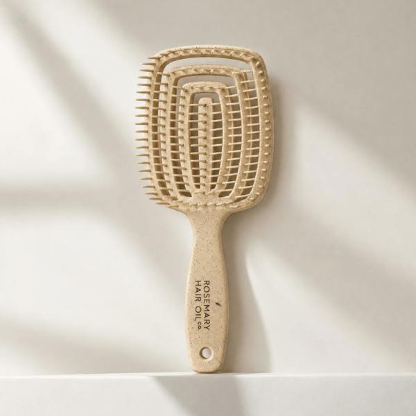 A natural hairbrush with a rectangular head and a long handle, standing upright on a white surface against a light beige background with soft diagonal shadows. The handle is printed with 'ROSEMARY HAIR OIL CO. Beige hairbrush with 'Rosemary Hair Oil Co.' branding on a beige background, detangle brush, tangle teaser, flexi brush