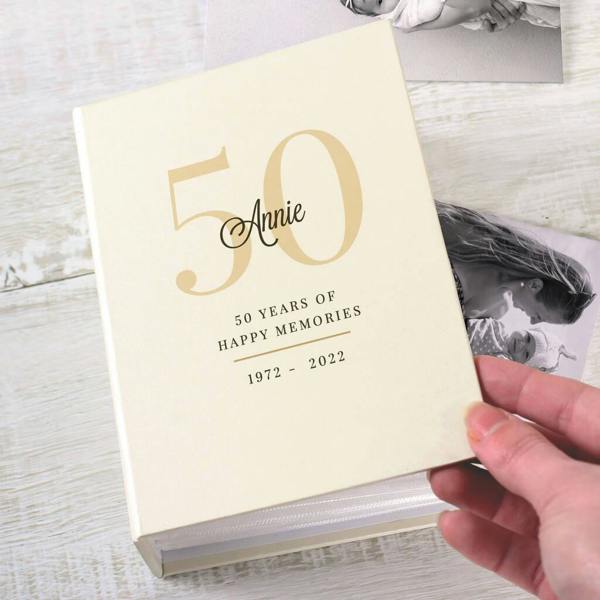 A close-up shot of a cream-coloured milestone birthday photo album held by a hand, featuring gold text that reads "50 Annie", "50 Years of Happy Memories", and "1972-2022", with blurred black and white photos visible in the background.