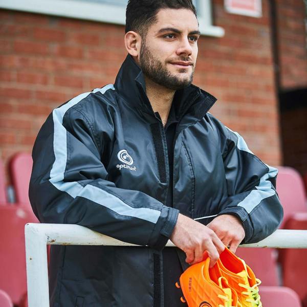 Man wearing black and grey Optimum sideline sub jacket holding orange boots, brick wall behind - Optimum