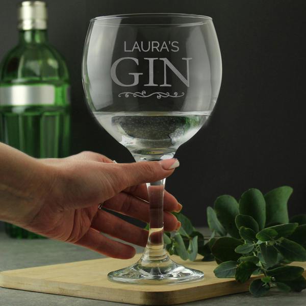 A person's hand holding a personalised stemmed gin balloon glass by Abode Home & Gifts, featuring the custom laser-engraved text "LAURA'S GIN" above a decorative scroll, filled with a clear drink on a wooden serving board next to a green gin bottle and leafy eucalyptus sprigs.